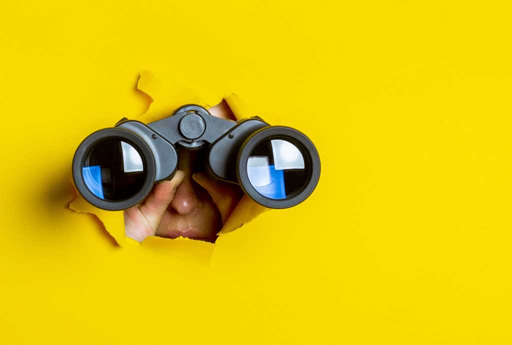 A person with binoculars peers through cracks in a yellow wall.