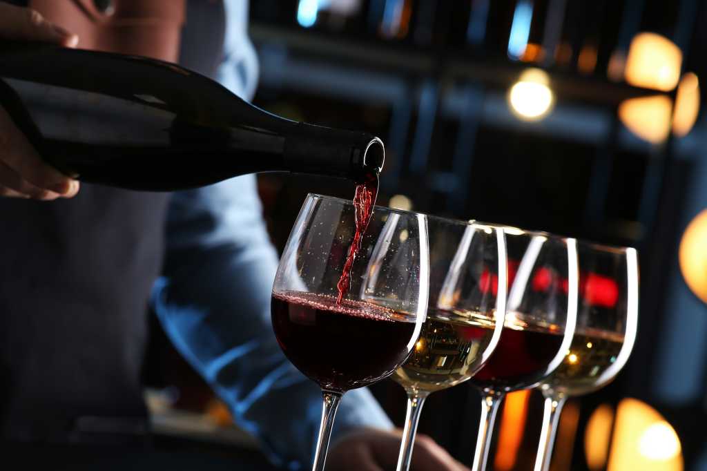 Server pouring red wine into four glasses against a dimly lit background