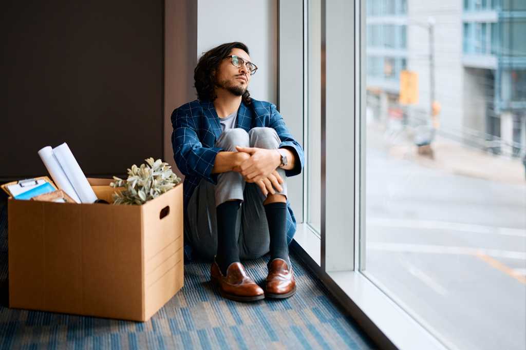 Sad entrepreneur sitting next to box of his belongings and looking through the window after being fired at work. Copy space.