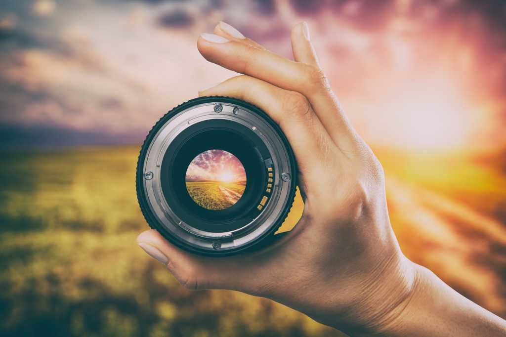 A hand holds a photographer's lens looking out at a field at sunrise. Preview, new, viewing the horizon.
